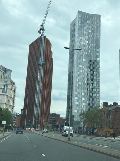 The image depicts a street scene in Manchester, United Kingdom, featuring several prominent buildings. The two most noticeable buildings are skyscrapers. One is clad in brown, horizontally-oriented panels, appearing to be under construction as indicated by the presence of a large crane attached to its upper portion. The second skyscraper is covered in a grid-like pattern of reflective windows with a section of different alternating patterns. A third building is visible on the roadside.
The street shows light vehicular traffic, including vans and cars. Pedestrians can be seen on the sidewalk next to the buildings. The sky appears overcast with a uniform cloud cover.
