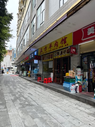 This is a street-level, eye-level, medium shot of a pedestrian walkway in front of shops on a city street in Liwan District, China. The walkway is paved with grey concrete tiles. On the right side of the walkway, there are several shops. One shop has a prominent yellow sign with red Chinese characters, which reads "正方羊肉馆" (Zhengfang Yangrouguan), indicating it is a mutton restaurant. This shop has a glass door and window display, showing pictures of food. In front of this shop, there are stacks of boxes, plastic crates, and large bottles of water. There are also three small red plastic stools. Next to this shop, to its right, is another shop with a red sign and a metal roll-up gate. The building above the shops has multiple stories with many windows and air conditioning units. On the left side of the walkway, there is a large tree with green foliage and a sidewalk beyond that, leading to other buildings. Two individuals are walking away from the camera on the left sidewalk. The sky is overcast.