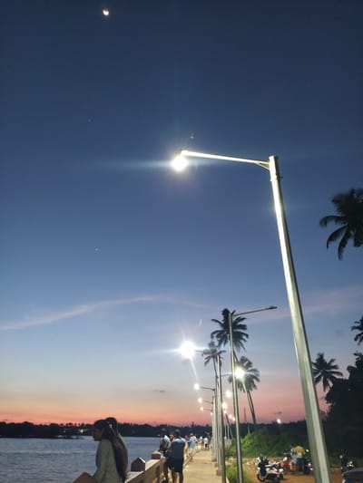 The image depicts a waterside scene at dusk. The sky transitions from a deep blue to a pale pink/orange hue on the horizon. Along the waterfront, a row of streetlights illuminate the walkway, which is lined with palm trees. People are standing or walking on the walkway near a low barrier that separates the walkway from the water. In the background, parked vehicles, including motorcycles, can be seen. A crescent moon is visible in the upper left of the image. The location is Badem, India.