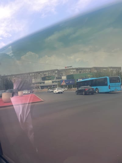 The image captures a street scene in the city of Pavlodar, Kazakhstan, likely on a cloudy day. In the foreground, there is a street with a traffic intersection. Several vehicles are visible, including a blue bus, a black sedan, and a white sedan. The bus is large and appears to be a public transport vehicle. The black sedan is positioned behind the bus, and the white sedan is to the right of the intersection.
In the background, there is a long, multi-story building, characteristic of Soviet-era architecture, with many windows. Several businesses and signs are visible on the facade of this building. One prominent sign displays "KMF" in red lettering. Another sign shows "ПЛАН" in gold lettering with stars above it, and further to the right, a sign reads "NEOMATOVA." There are also trees and some greenery visible around the buildings.
Reflections on the surface from which the photo was taken are noticeable in the upper and left portions of the image, creating a slightly distorted or layered effect, particularly on the sky and the foreground elements. The sky is filled with clouds, with patches of lighter blue sky visible. The overall impression is a typical urban environment with vehicular traffic and commercial establishments.