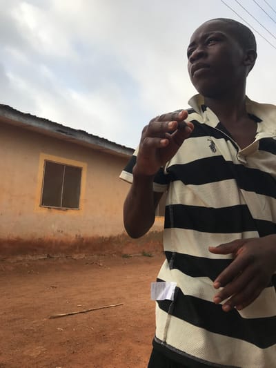 The image displays a young person with dark skin and short, dark hair, standing outdoors during daytime. The individual is wearing a black and white striped polo shirt and is looking downwards and slightly to their left with a neutral expression, holding their hands loosely in front.

In the background, to the person's right, there is a building with an earth-toned, possibly weathered, wall. A simple, dark-framed window with a grid pattern is visible on the building's facade. Above the building, a light, overcast sky suggests a cloudy day. The ground in the lower right portion of the image is reddish-brown earth, with a thin stick lying upon it. This outdoor setting, characterized by the earthy ground and the building style, is consistent with a location such as Ilorin, Nigeria.

Notable details include a small, embroidered emblem, resembling a polo player logo, on the left chest of the person's shirt. A white garment tag with illegible text, appearing to be in an Asian script with some numbers, hangs from the bottom hem of the shirt. The scene captures a casual, everyday moment in an outdoor environment.