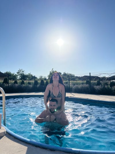 A man and a woman are in a swimming pool. The woman is on the man's shoulders, and both are smiling. They are in a pool with clear blue water, surrounded by a concrete deck. In the background, there is a fence, trees, and buildings. The sun is shining brightly, casting reflections on the water. The sky is a clear blue. This appears to be a sunny day, likely in the late morning or early afternoon, judging by the angle of the sun and the bright light. The presence of the swimming pool, the sun, and the light clothing suggest a warm weather activity. The scene is set in Lozen, Bulgaria. No visible text is present in the image.