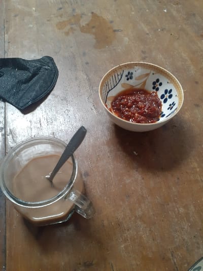 The scene is set on a wooden surface, likely a table, within the city of Cimenyan, Indonesia. The surface exhibits visible wood grain, wear marks, scratches, and several dried or semi-dried liquid stains. In the upper left quadrant, a black fabric face mask is positioned, appearing folded or crumpled. In the upper right quadrant, a small, light-colored ceramic bowl with blue and brown floral patterns contains a quantity of reddish-orange chili paste, possibly sambal, with visible coarse chili fragments. In the lower left quadrant, a clear glass mug holds a light brown liquid, consistent with coffee or a similar beverage, with a silver-colored metal spoon resting vertically within it. No individuals are present in the image.