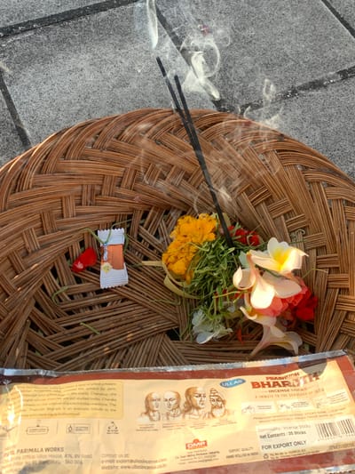 This is a close-up, eye-level shot of a woven basket placed on a textured grey surface that appears to be asphalt or concrete. Two dark incense sticks are burning, emitting wisps of white smoke that curl upwards. Within the basket, there is a small arrangement of flowers, predominantly yellow and white, with some green foliage and a single red petal. In the lower portion of the image, a package of "PRABHUDH BHARATH INCENSE STICKS" is visible. The package displays text and illustrations of historical figures. A small, white and orange foil-wrapped object, possibly a charm or token, is also present within the basket, attached by a green string. The scene is outdoors, suggested by the lighting and the textured ground. There are no people or animals visible in the image.