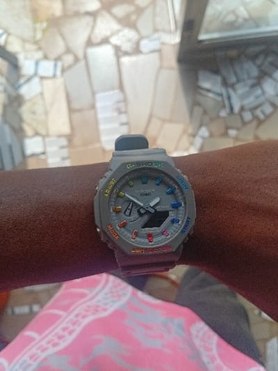 A dark-skinned human forearm, oriented horizontally, displays a grey Casio G-Shock GA-2100 series watch with a grey resin band. The watch face features a combination analog-digital display, multicolored hour markers, and white operational text including "ADJUST," "MODE," "PROTECTION," "START," and "LIGHT." Below the forearm, a segment of pink fabric patterned with white and grey geometric or abstract designs is visible. The background consists of an outdoor paved surface comprised of irregular, light-toned mosaic-style tiles, possibly a patio or walkway. A blurred, reflective surface, potentially glass, is present in the upper right quadrant, partially obscuring additional tiling. The scene is illuminated by ambient daylight. The location is Ifite-Awka, Nigeria.