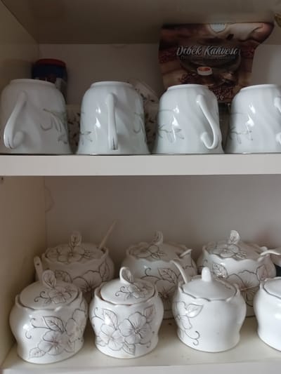 The image shows a shelf with white ceramic tea cups and sugar bowls, decorated with a floral pattern. The cups are on the top shelf, and the sugar bowls are on the bottom shelf. The background is a plain, light-colored wall. The scene appears to be indoors, possibly in a kitchen or dining room. There are no people or animals visible in the image. The lighting is even, suggesting an indoor environment with artificial light. The overall impression is one of a well-organized collection of tableware.