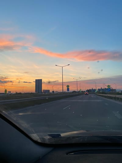 Captured through a car window at sunset, the image shows a highway receding into the distance. A car with red taillights is visible on the road. Adjacent to the highway, a series of tall, cylindrical structures are present; these are storage silos. Streetlights line the roadway. Speed limit signs are also visible. The sky transitions from blue to orange and pink. The location is Stroevo, Bulgaria.