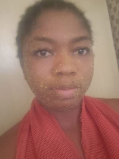 A front-facing, waist-up shot depicts a young individual with dark skin and dark, textured hair, looking directly into the camera. Their face is covered with a granular, yellowish-white substance, applied unevenly, particularly visible on the forehead, cheeks, nose, and chin. The individual's eyes are open, and their lips are slightly parted. A red, open-weave, mesh-like fabric, possibly a towel or garment, is draped around their neck and shoulders. The background is out of focus, appearing as a plain, light-colored surface on the left and a subtly textured, lighter surface on the right. There are no visual cues or identifying markers within the image to indicate the specific geographical location as the city of Kakau, Nigeria, or any other specific region.