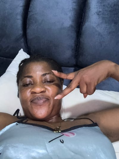 A woman is lying on a bed, possibly in Ajara, Nigeria, and taking a selfie. She is on her back with her head resting on a white pillow against a dark blue, tufted headboard. The woman has dark skin, braided hair, and is wearing a light blue camisole. She is smiling and holding up her right hand in a peace sign. Her nails are painted a light peachy color. The lighting appears to be indoor, and the overall mood is casual and relaxed.