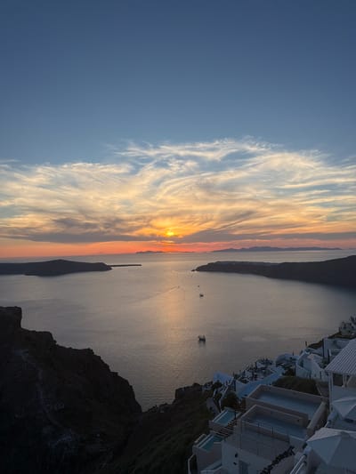 A sunset scene overlooks the caldera in Santorini, Greece. The sun, a bright orange disc, is setting on the horizon, casting a warm glow across the sky and water. Wispy clouds streak across the sky, tinged with shades of orange, pink, and blue. The caldera, a large volcanic crater filled with water, dominates the middle ground of the image. Islands, silhouetted against the sunset, are visible in the distance. The foreground features a cliffside with a village of white buildings cascading down its slope. Some of the buildings have private pools. Boats are scattered across the water.