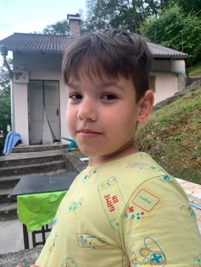 This is a close-up, vertically oriented photograph of a young boy wearing a yellow t-shirt adorned with a pattern of video game controllers and consoles. The shirt has text that reads "GAME ON" and "loading...". The boy's face is partially visible, with his right eye looking directly at the camera. He has dark hair and light skin. The background is blurred but suggests an outdoor setting with greenery and what appears to be a building with a gray roof and a white wall. Steps are also visible, possibly leading up to the building. The lighting suggests it is daytime. The overall mood appears casual and relaxed. The location context provided is Novo bardo, Bulgaria.
