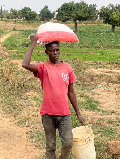 In an outdoor setting, a man stands in a field in Udu, Nigeria. He carries a white and red sack on his left shoulder and holds a wire attached to a beige plastic bucket with his right hand. He wears a red T-shirt and dark pants. The field consists of dry dirt and patches of green grass. Trees are visible in the background.