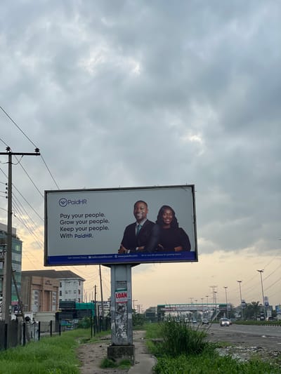 The image captures a billboard advertisement for PaidHR, positioned on a roadside in Ojomu, Nigeria, under a cloudy sky. The billboard features a logo for PaidHR and a tagline reading "Pay your people. Grow your people. Keep your people. With PaidHR." Two individuals, a man and a woman, are pictured on the billboard, both smiling and appearing professional. Below the main text, smaller print indicates "Get Started Today," an email address "hello@paidhr.com," and a website "www.paidhr.com," along with the descriptor "All-in-one HR software for growing teams."
The billboard is mounted on a concrete pillar. To the left of the billboard, a tall utility pole with numerous wires is visible, along with a multi-story building. In the foreground, a grassy embankment lines the side of a dirt and rubble-strewn path. Beyond the billboard, a highway with several cars and streetlights can be seen, extending into the distance with more utility poles and structures. A pedestrian bridge with green and white signage is also visible further down the highway. The overall environment suggests an urban or suburban setting in Nigeria.