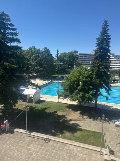 The image shows an outdoor swimming pool complex in Sandanski, Bulgaria. The pool is surrounded by greenery, including tall trees and grass areas. A multi-story building is visible in the background. A white tent-like structure is located near the pool, with possible tables underneath. A lamppost stands in the foreground on a paved area. A group of people is visible near the bottom of the frame.