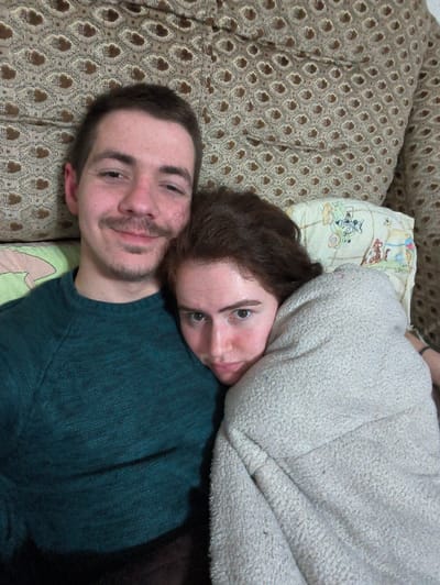 An indoor close-up photograph depicts two individuals, a male and a female, in the city of Negorci, North Macedonia. The male, on the left, has short dark hair, a mustache, and stubble, wearing a dark teal long-sleeved sweater, and looks directly at the camera with a faint smile. The female, positioned to the right, has reddish-brown hair and is leaning her head against the male's left shoulder. She is wrapped in a light-colored, textured blanket and looks towards the camera with a neutral expression. A light-colored pillow with a multi-colored cartoon pattern is visible behind the female's head. The background consists of a light brown, patterned upholstery fabric.
