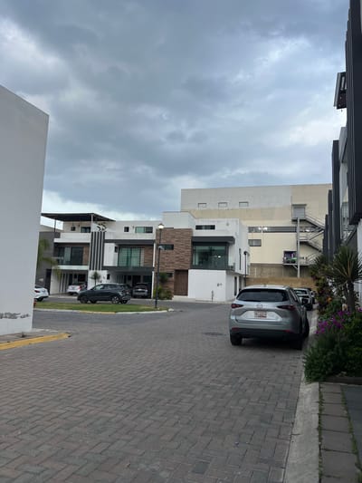 The image depicts a street scene in Cholula, Mexico, characterized by modern multi-story townhouses with predominantly white facades accented with brown and black architectural details. Several vehicles are parked along the brick-paved street, including a silver Mazda SUV with a license plate visible on the rear. The sky is overcast with gray clouds, suggesting a cloudy day. A commercial building with external stairs is visible in the background. The environment includes landscaping with trees, shrubs, and flowers along the edge of the street. The scene is residential, with a clear focus on the built environment.