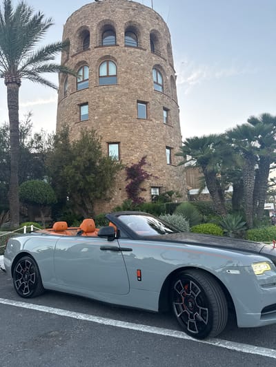 The image shows a grey convertible Rolls-Royce parked next to a rustic stone tower in Marbella, Spain. The tower has multiple rounded windows and appears to be a historical building. Lush greenery, including palm trees and bushes, surrounds the tower and the car, suggesting a warm climate. The car's interior is visible and features orange leather seats. The scene is set outdoors during daylight, possibly late afternoon or early evening, given the soft lighting. There is no discernible activity or event taking place; it appears to be a static shot of the car and its surroundings. The sky is light blue with some wispy clouds. The image is oriented vertically, with the tower on the left and the car on the right. No visible text is present in the image.