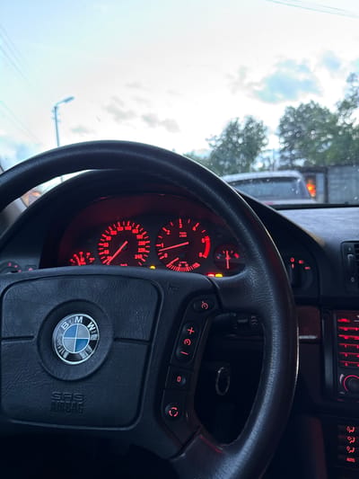 The image shows the interior of a BMW car, specifically the steering wheel and dashboard. The steering wheel has the BMW logo and airbag inscription. The dashboard features red-lit gauges and climate control buttons. The car appears to be stationary, with a view of trees and sky visible through the windshield. Location cannot be determined.