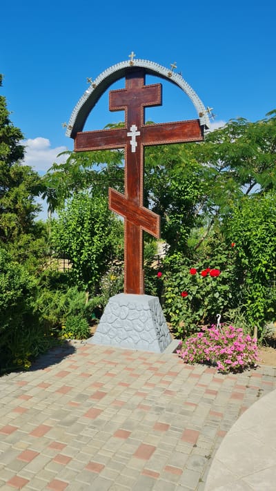 This is a medium shot of a large, wooden Orthodox cross adorned with a silver, semi-circular halo, standing outdoors in a garden. The cross is the central subject, its dark brown wood contrasting with the bright blue sky behind it. Smaller golden crosses are affixed to the halo. The cross itself has a white decorative element on its horizontal bar.

The setting is a lush garden with vibrant green foliage, including trees and bushes. Red roses bloom near the cross, and a pot of pink flowers hangs to the right. The ground is paved with interlocking bricks in shades of beige and pink, forming a walkway. A grey stone pedestal with a textured pattern sits to the right of the cross. The scene is well-lit, suggesting a sunny day.

The image captures a serene and possibly sacred atmosphere, evoking a sense of peace and reverence. The orientation of the image is vertical. There is no visible text in the image. Given the location context of Bilhorod-Dnistrovskyi, Ukraine, this may be a religious site or monument within the region.