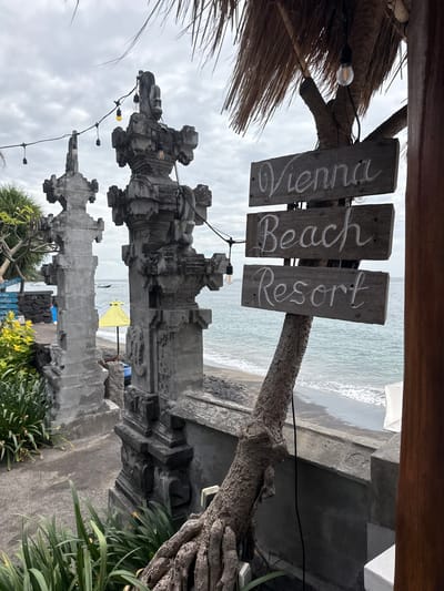 The image depicts the entrance to the "Vienna Beach Resort" in Abang, Indonesia. Three wooden signs, hand-painted with white lettering, are mounted on a textured tree trunk. The signs read "Vienna," "Beach," and "Resort." In the background, the ocean is visible with waves breaking on a dark sand beach. Two large, ornate stone pillars flank the pathway, suggesting an architectural entrance. String lights with exposed bulbs hang between the pillars and above the signs, indicating an evening or overcast setting. Lush green plants grow in the foreground and around the base of the pillars. The overall atmosphere suggests a tropical beach resort setting with a focus on natural elements and a touch of rustic charm.