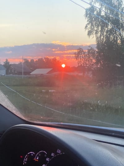 The image depicts a sunset viewed from inside a vehicle, looking through the windshield. The sun, a bright red orb, is setting over a rural landscape. In the foreground, part of a car's dashboard is visible, including a speedometer and fuel gauge. Beyond the dashboard, a field with grass and some vegetation stretches towards a line of trees. Several houses with roofs are situated in the mid-ground, suggesting a small village or settlement. The sky is filled with hues of orange, pink, and purple, with some scattered clouds. The overall impression is that of a peaceful rural sunset. The location is identified as Sukhaia Balka, Russia.