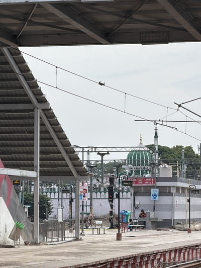 The image depicts a scene at a train station in Lucknow, India. In the foreground, a concrete platform with a red and white striped border is visible, leading towards a covered waiting area with a sloped, corrugated metal roof supported by metal beams. A green trash can and a metal bench are placed on the platform. In the middle ground, a building with a patterned facade houses a public restroom facility, with separate signs indicating "LADIES" and "GENTS" restrooms. A man is seated near a water fountain outside the restroom. Further back, a large green dome with white detailing, characteristic of Islamic architecture, is prominently featured, likely belonging to a mosque. A minaret also rises above the trees. Overhead, a complex network of railway catenary wires is suspended. The sky is overcast. In the background, the ornate facade of a building with decorative white fencing can be seen. The overall scene suggests a busy public transportation hub with elements of local religious architecture.