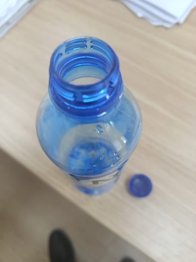A blue plastic water bottle with a partially filled interior of clear liquid sits on a light brown wooden surface. The bottle is open, with the blue cap lying to the right of the bottle. In the upper left corner of the image, a stack of white papers is visible. The scene appears to be indoors, on a desk or table.