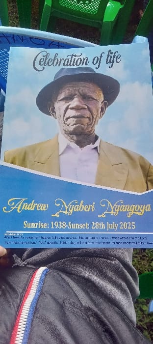 The image depicts a "Celebration of life" program pamphlet, held by an individual, in an outdoor setting in Esise ward, Kenya. The program features a central portrait of an elderly man identified as Andrew Nyaberi Nyangoya. He is shown wearing a dark hat, a light-colored jacket, and a white shirt against a light blue background. Below the portrait, the text states "Sunrise: 1938-Sunset: 28th July 2025," followed by a partially visible quote, likely a biblical verse. The lower portion of the image shows the lap of the person holding the program, clad in dark grey or black sweatpants with a vertical red, white, and blue striped accent on the left side. In the blurred background, green plastic chairs and green ground cover, possibly grass, are visible, indicating an outdoor gathering.