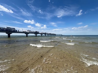 This is a photograph of the pier in Międzyzdroje, Poland. The pier extends far out into the Baltic Sea, with several small buildings and kiosks situated along its length. The sky is a vibrant blue, dotted with white, wispy clouds. Gentle waves lap at the sandy shore in the foreground, where a dark rope is visible lying on the seabed. In the distance, a small red boat can be seen on the water, and a few people are swimming or wading near the shore. The overall scene conveys a sense of a bright, sunny day at the seaside.
