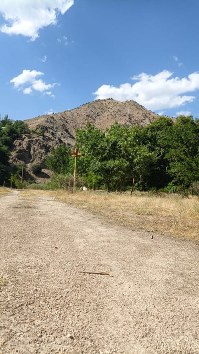 The image depicts a dirt road leading towards a rocky, mountainous landscape under a bright blue sky with scattered white clouds. A rusted metal post with a red crossbar stands on the right side of the road, amidst dry grass and green trees. The terrain is arid, with sparse vegetation on the mountainside. The scene appears to be a rural or natural environment, potentially in or near the city of Mirat, Tajikistan.