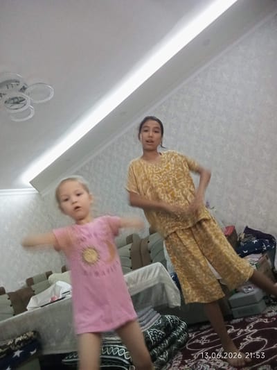 This is a low-angle, indoor, full-body photograph featuring two young females. The female on the left, with blonde hair, is wearing a pink t-shirt with a yellow sun graphic and blue shorts. She is in motion, with her arms outstretched to her sides. The female on the right has dark hair and is wearing a yellow patterned outfit consisting of a short-sleeved top and wide-leg pants. She is also in motion, with her hands near her waist. Both are on a patterned rug. In the background, there is furniture, including sofas and a table covered with a white cloth. The room has a textured wallpaper and a modern ceiling light fixture with multiple rings. A timestamp in the lower right corner indicates "13.06.2026 21:53". The image was likely taken in a residential setting in the city of Bekabad, Uzbekistan.