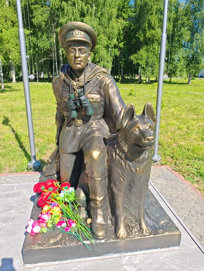 This is a bronze statue of a soldier kneeling next to a German Shepherd dog. The soldier is wearing a military uniform with a hat, binoculars around his neck, and what appears to be a rifle slung over his shoulder. The dog is standing attentively beside him. In front of the statue, a bouquet of artificial flowers, including red, pink, and yellow blooms, is placed on the base.
The statue is located outdoors in a grassy park with trees in the background. Two tall, silver poles, likely flagpoles, are visible on either side of the statue. The ground around the base of the statue is paved with stone tiles. The lighting suggests it is a sunny day, casting shadows from the poles. The overall scene appears to be a memorial or monument.
There is no visible text in the image. The atmosphere conveyed by the statue is one of solemnity and companionship, with the soldier and his dog depicted in a moment of watchful readiness. The presence of the flowers suggests a place of remembrance and tribute. The location is identified as Buy, Russia.