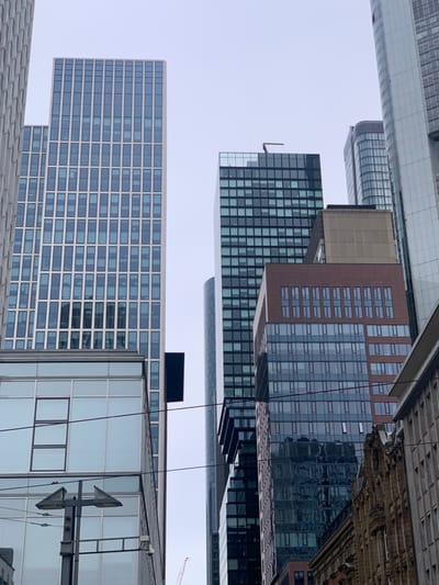 The image shows a view of several office buildings in Frankfurt am Main, Germany. The location is identifiable as the city's financial district, known as the Bankenviertel, based on the coordinates provided. Several tall buildings with glass facades dominate the scene. On the left, there are two buildings: one with a light-colored exterior and a more modern design, and another with a darker exterior and a more traditional design. In the center, a taller building with black frames and dark glass dominates the view. To the right, there are several other office buildings, one with a brown facade and another with a more modern, sleek design. Power lines and a streetlight are visible in the foreground. The scene appears to be taken during the day, with a clear sky and minimal pedestrian activity visible.