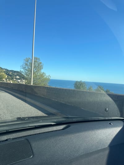 The image presents a daytime view from within a vehicle, looking forward over a coastal scene in Cap-d'Ail, France. The foreground displays a dark gray textured vehicle dashboard, featuring a speaker grille on the left and the base of the windshield with wipers. Light reflections are visible on the windshield's interior surface. Outside the vehicle, an asphalt road curves to the left, bordered by a light-colored barrier and a stone retaining wall. A tall, slender, light-colored pole ascends vertically from the lower-left, extending into the clear blue sky. To the left of the road, a vegetated hillside with scattered residential buildings is visible. To the right, beyond the retaining wall, the deep blue Mediterranean Sea stretches towards the horizon beneath a cloudless, bright blue sky. Scattered green trees are present on the hillside and partially visible above the retaining wall. The scene is uniformly illuminated by direct sunlight, casting distinct shadows across the vehicle's interior. No individuals or specific activities are observed.