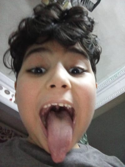 The image is a selfie taken by a young boy with short, dark, curly hair. He is wearing a grey t-shirt and is looking directly at the camera. His mouth is open and his tongue is sticking out. His teeth are visible and there appears to be a missing tooth. He is in what appears to be a room with a white ceiling. The location of the image is likely in the city of Alexandria, Egypt, based on the coordinates 31.227 latitude and 29.962 longitude. The image is likely taken indoors, based on the white ceiling and the wall in the background.