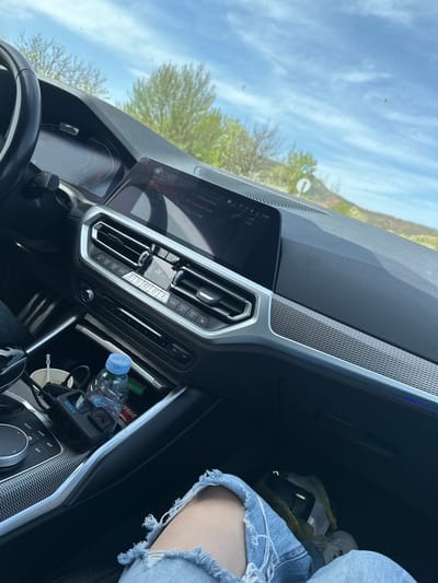 This media file captures the interior of a modern car, likely a BMW, from a passenger's perspective, looking towards the dashboard and out the front windshield. The scene appears to be set in Madrets, Bulgaria, during a bright, clear day with blue skies and scattered white clouds.
On the right side of the image, a person's leg, clad in distressed light-wash denim jeans, is visible, suggesting a relaxed passenger. The car's dashboard features a prominent infotainment screen displaying "11:45", indicating late morning. The screen also shows "Music collection", "Phone", "on Raycho", "Previous track", "Unknown track", "Delete track", "Sound", and "Artists". Below the screen, the climate control panel includes buttons for "AUTO", "MAX A/C", fan speed, temperature, and seat heating.
The central console houses an iDrive controller with surrounding buttons labeled "MENU", "MEDIA", "HOME", "MAP", "NAV", "BACK", and "OPTION". Visible in the storage compartment near the gear shifter are a blue-capped water bottle, a black GoPro camera (GoPro Hero 9 Black, recognizable by its design and branding), and a small white cup. A red "VIVA" snack wrapper is also visible. In the footwell or glove compartment area, a yellow plastic bag containing a white styrofoam container and a dark-colored object (possibly a phone or wallet) is present.
Through the windshield, green trees with spring foliage and a partial view of a road sign (a circular sign with a diagonal line) are visible. The driver's instrument cluster shows "63" (likely speed) and "D" (drive mode), along with "-16.5", which could be an outside temperature reading. The overall impression is of a casual car journey under good weather conditions.