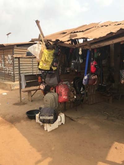 The image displays an outdoor market stall located in Ogbese, Nigeria. The stall features a rustic construction with rough wooden beams and posts supporting a worn, tan-colored corrugated metal roof exhibiting visible damage. Hanging from the wooden framework are numerous bags and backpacks of varying sizes and designs, including yellow, red (with "SINCE 1990" visible), grey, black, blue, and patterned items, some still in clear plastic wrapping.

On the reddish-brown, dusty ground in front of the stall, a grey backpack is positioned atop two white concrete blocks, alongside a dark blue messenger bag. A black plastic basin rests on the ground nearby. To the left of the main stall, an adjacent building with corrugated siding or horizontal wooden planks has a simple wooden bench affixed to its exterior, draped with a light-colored fabric. A single bare light bulb hangs from the interior roof structure of the stall. In the upper left background, a street light pole equipped with a solar panel is discernible against an overcast sky. No individuals are present in the scene, and no actions or interactions are depicted.