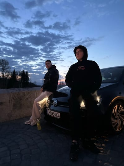 The image displays two males at dusk in Nevestino, Bulgaria, positioned on a paved area with cobblestones. A black Volkswagen Golf is centrally located, facing slightly left. The vehicle exhibits a Bulgarian license plate "KH 8954 KA" and "GTD" badging on its grille.
On the left, a male with a short, dark haircut wears a dark patterned puffer jacket, light-colored wide-leg pants, and white athletic shoes. He is seated on a low stone barrier or wall, with his right leg bent and resting on the car's hood. A yellow plastic bottle is visible on the ground near his feet. His gaze is directed towards the viewer.
On the right, a second male, wearing clear-framed glasses, a black hooded sweatshirt with a red graphic, dark pants, and black shoes, stands leaning against the front passenger side of the car. He looks slightly to his right, away from the viewer.
The background features a natural landscape of hills and trees, silhouetted against a sky showing a gradient from deep blue at the top to shades of orange and pink near the horizon, indicative of twilight. Scattered clouds are present across the sky.