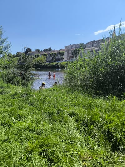The image shows a scene in Chézenas, France, on a sunny day. In the foreground is lush green grass, obscuring the view of the water. In the water, there are several people. Some appear to be swimming, while others are standing and interacting in the water. One person is wearing a yellow hat. In the background, there are buildings of varying heights and architectural styles, lined up along a hill. Trees are interspersed among the buildings. Vegetation is present along the shoreline, separating the water from the built environment. A church steeple is visible to the right of the image.
