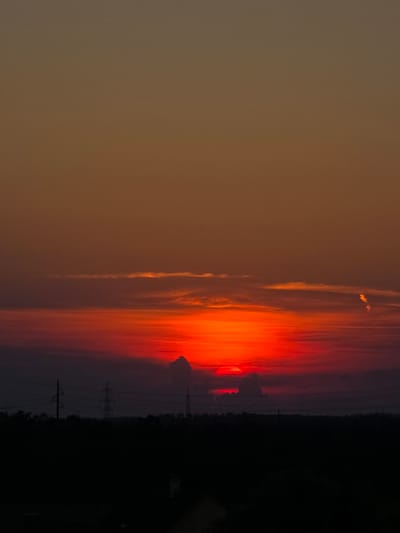 A dramatic sunset paints the sky over Bruck an der Leitha, Austria, with vibrant hues of orange and red. The sun, a brilliant disc, is partially obscured by dark, silhouetted clouds, creating a stark contrast against the fiery sky. Wisps of lighter clouds stretch horizontally across the upper half of the frame, catching the last rays of sunlight.

Below the celestial display, the landscape is cloaked in deep shadow. The dark silhouette of a treeline forms a dense border across the lower part of the image. Emerging from this darkness are the skeletal forms of electricity pylons, their towering structures adding a touch of industrial modernity to the natural spectacle. The overall scene is one of dramatic natural beauty, tinged with the imposing presence of human infrastructure.