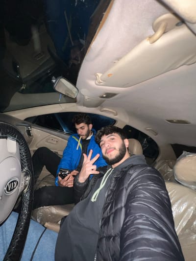Two men are inside a Kia car. One man in a black puffer jacket makes a peace sign. Behind him sits another man in a blue jacket, holding a cigarette. The interior of the car is visible, including the steering wheel, sun visor, rearview mirror, and seats covered with plastic. The photo appears to be taken at night.