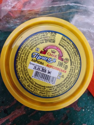 This is a close-up, overhead shot of a yellow plastic tub of mustard, likely in a food preparation setting. The tub has a blue and yellow label with Ukrainian text and a barcode. The label indicates the product is "Gorchitsa Korolivska" (Crown Mustard) and weighs 100 grams. It also provides nutritional information and an expiration date of 28.09.2026. The tub is placed on a dark surface with some red and green streaks, possibly a table or countertop. The background is blurred, showing plastic packaging. The lighting appears to be artificial, casting a slight shadow from the tub. The overall impression is of a product ready for use or storage.