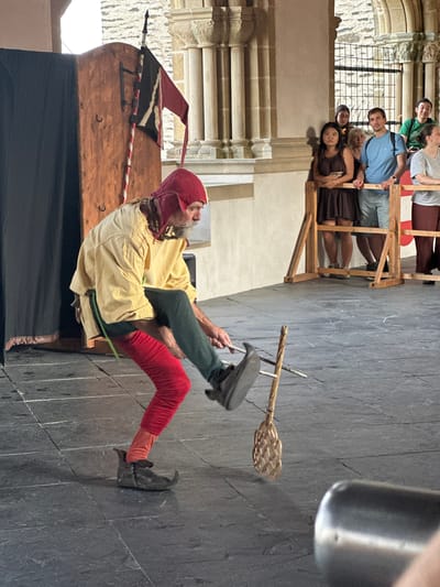 In front of the Vianden Castle in Vianden, Luxembourg, a performer in historical costume is demonstrating a skill. The performer, wearing a red hood, a yellow tunic, green leggings, and red stockings, is mid-motion with his right leg extended, kicking a woven object attached to a stick. The object is a basket-like tool, possibly for a demonstration of historical entertainment or skill. Behind the performer, a wooden structure with a black curtain stands to the left, and a historical flag with red, black, and white stripes is mounted on a pole to the right of the wooden structure. Spectators are visible in the background, standing behind a wooden barrier, observing the performance. The ground is paved with large, dark stone tiles. The architecture in the background suggests the historical setting of the castle.