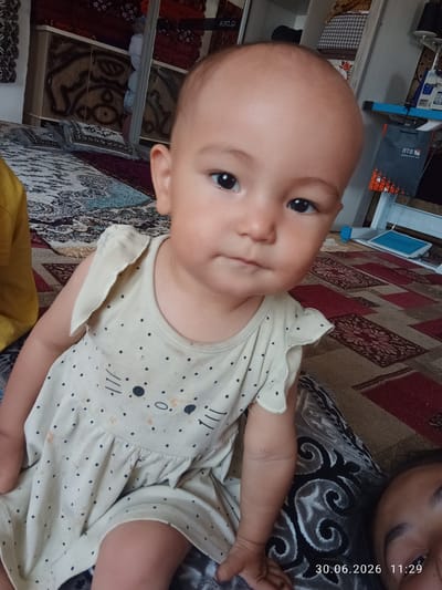 In a residential setting in Batken City, Kyrgyzstan, a close-up portrait shows a young child, likely a toddler, seated. The child is wearing a light-colored, sleeveless dress adorned with a pattern of small black dots and a graphic of a cat's face. The child's head is tilted slightly, and they are looking directly at the camera with dark eyes. The background is a patterned rug and a blurred view of furniture. A portion of another person's face is visible in the lower right corner of the frame. The date stamp "30.06.2026 11:29" is overlaid on the image.