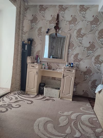The image depicts an indoor scene, likely a bedroom or dressing area, in Komyshivka, Ukraine. A vanity table with a mirror stands against a wall covered in floral wallpaper. Various cosmetic items and a handbag are arranged on the table. To the left of the vanity is a tall, dark cabinet. The floor is covered with a patterned rug. A stuffed toy monkey hangs from a string attached to the wall above the mirror. The lighting suggests it is daytime, with natural light coming from an unseen window.