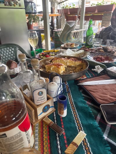 The image captures a cluttered outdoor table scene, likely from a gathering. The location is Elena, Bulgaria as evidenced by "Ouzo Plomari", a spirit from the Greek island Lesbos, and the traditional woven rug underneath.

On the table, a variety of items are present. There's food in a silver pan resembling a pie or baked dish, along with other plates of food. Several alcoholic beverages are scattered around, including bottles labeled "Ouzo Plomari Isidoros Arvanito", a green beer bottle ("Tuborg Copenhagen"), and a bottle with "RECYCLE ME" printed on its label. Glasses, some with liquid inside, are also visible. 

Other objects include what appears to be a wooden holder for small liquor bottles, possibly the "Ouzo Plomari", a phone, a brown cigar and a knife. The table is covered with a traditional Bulgarian Kilim rug.

Beyond the table, a glimpse of the background reveals what appears to be an outdoor setting with a drying rack. The scene seems to depict the aftermath of a meal or social gathering.