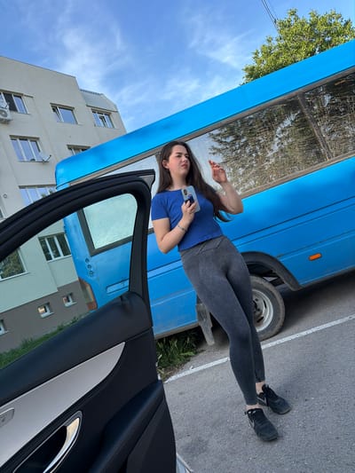 A young woman with long dark hair is standing next to an open car door, holding a smartphone in her left hand. She is wearing a blue t-shirt and grey leggings. Behind her is a blue bus and a light-colored building. The scene appears to be a street in Kaspichan, Bulgaria, under a cloudy sky.