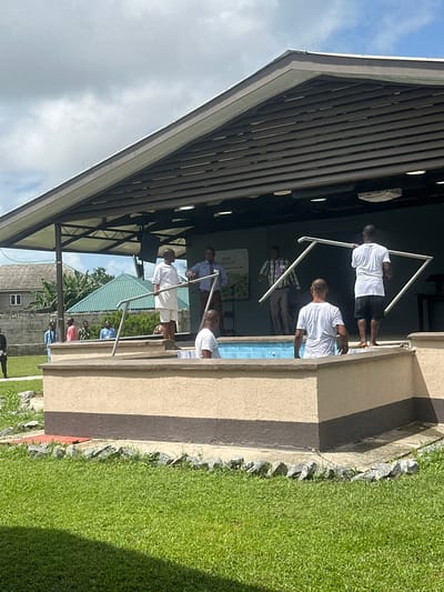 In Itoga, Nigeria, outdoors, on a bright day with scattered clouds, a baptism is taking place in a raised, tiled pool. The pool's exterior is a light tan with a dark brown band around its base. A group of men are present.
In the pool, a man wearing a white t-shirt and black shorts stands with his back to the camera. In front of him, a man in a white robe and shorts is partially submerged. To his right, another man in a white t-shirt and black shorts is standing, facing away from the camera and holding a white frame. Another white frame is being held by a man further back, who is wearing a white t-shirt and black shorts, facing away from the camera.
Behind the pool, on a raised platform, two men are talking. One is wearing a light blue shirt and dark pants, and the other is in a checkered shirt and grey pants. Behind them, a large green sign with text in white and green is visible.
To the left of the pool, several other men are standing. One man in a white t-shirt and light shorts stands on the edge of the pool. Another man in a light blue shirt and black pants stands behind him. Further to the left, a man in a dark suit stands on the grass.
In the background, a building with a brown, slatted roof extends over the platform. Houses with green roofs and a tall tree are visible behind the building. The area around the pool is covered in lush green grass with scattered grey rocks.