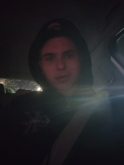 The image captures a male individual seated in the passenger position of a vehicle, located in Wemmel, Belgium, during low-light conditions. The subject, a young adult, has short dark hair and wears a dark hooded sweatshirt with the hood up, covering a backwards-worn baseball cap displaying a brown/orange underside of the brim. An inverted "Calvin Klein" logo is visible on the chest of the sweatshirt. A seatbelt diagonally crosses the individual's torso. Through the vehicle's rear window, blurred light sources and apparent precipitation or condensation on the glass are discernible. A prominent diagonal light flare extends across the lower right portion of the frame.