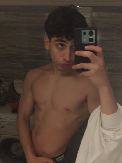 A shirtless young man takes a selfie in a bathroom. He has dark curly hair and is holding a smartphone with a multi-lens camera. His physique is visible, showing defined chest muscles. Part of a washing machine and toiletries are visible in the background. He has a white garment draped over one arm. The bathroom walls have a textured, gray-toned surface. The lighting in the room appears to be dim, and the overall image has a slightly grainy quality.